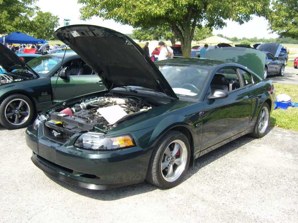 Dark green Ford Mustang, hood open, parked next to another green Mustang. Other cars and tents in background