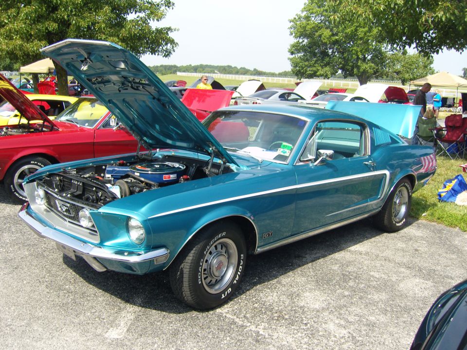 Teal 1967 Ford Mustang fastback with hood and trunk open, parked on asphalt with other cars in background