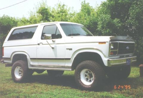 1995 Ford Bronco - This 1980s Ford Bronco is shown from the front three-quarter view, with a grassy area and trees in the background.
