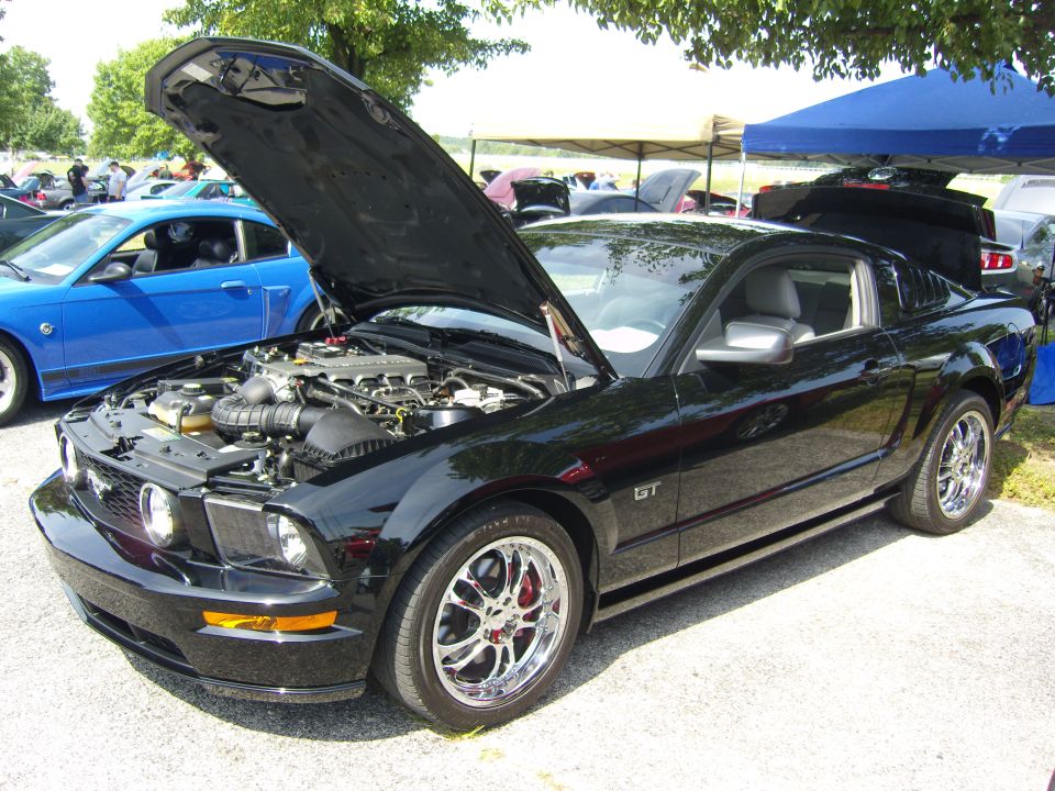 Black Ford Mustang GT with hood open, chrome wheels, and red brake calipers. A blue Mustang is visible in the background