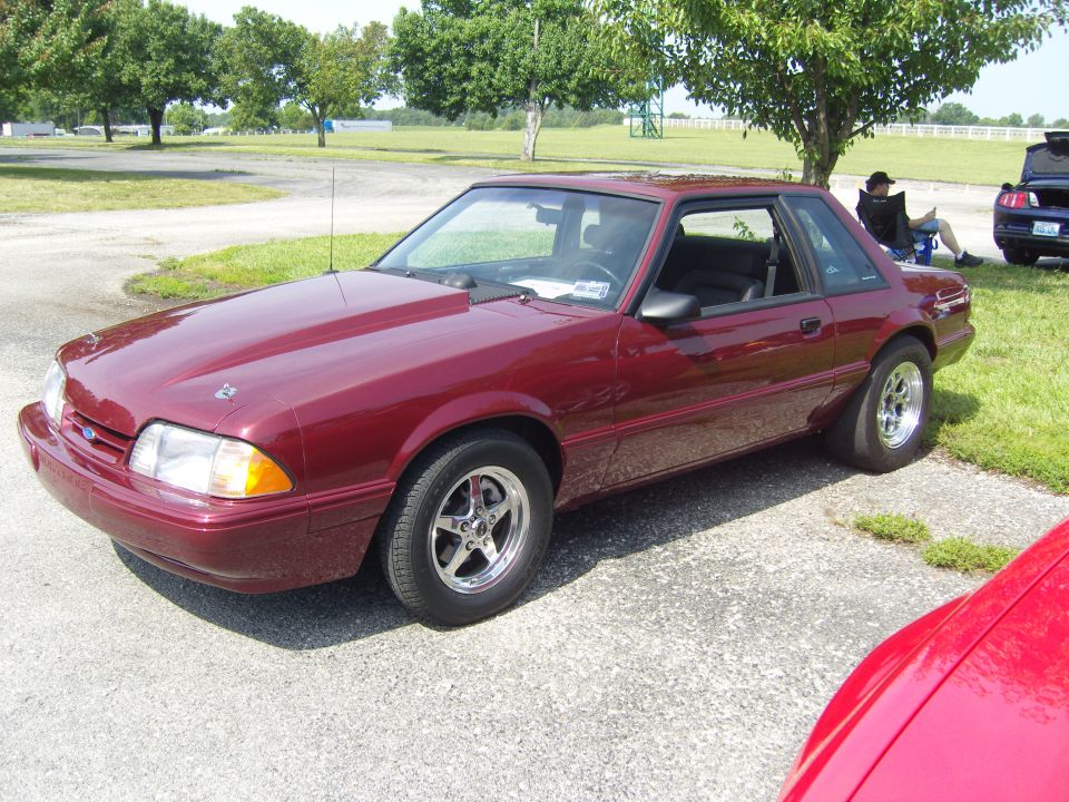 Maroon Ford Mustang coupe with aftermarket hood and drag tires, parked outdoors. Another car is partially visible in the foreground