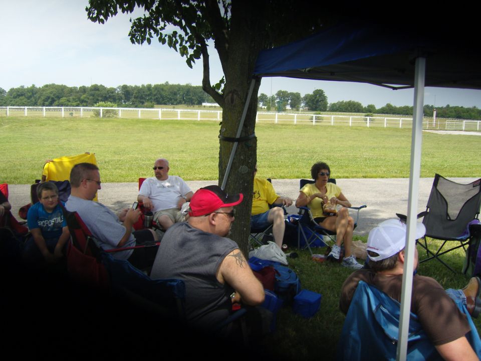 People sit in lawn chairs under a blue canopy, with a grassy field and white fence in the background