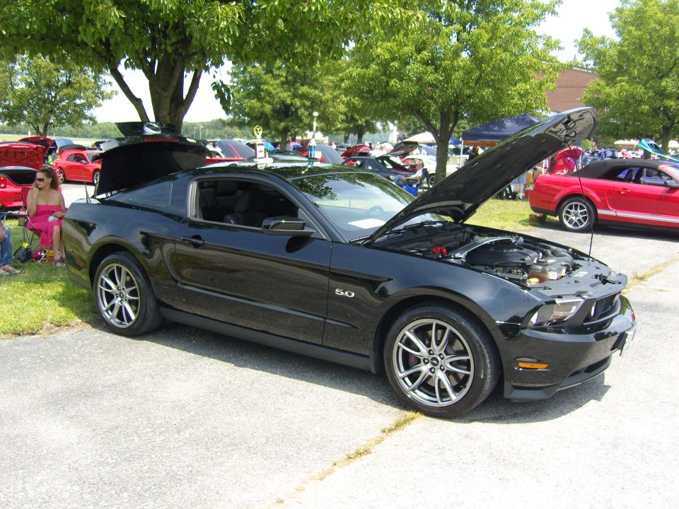Black Ford Mustang parked outside with hood open, showing engine. Another red convertible Mustang is visible in the background