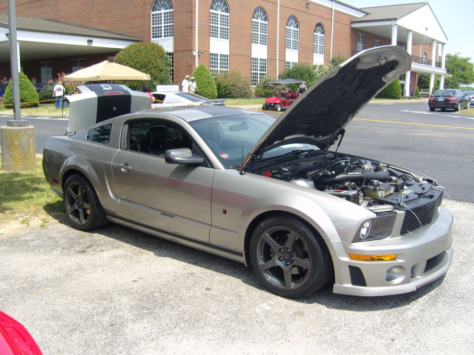 Silver Ford Mustang with hood open, showing engine. Car has racing stripes on the hood and trunk