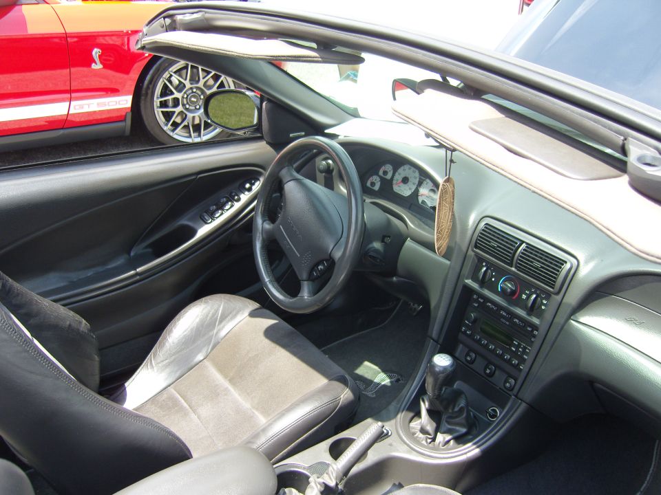 Interior of a 1999 Ford Mustang convertible showing dashboard, seats, and gear shift. Red Mustang GT 500 visible outside