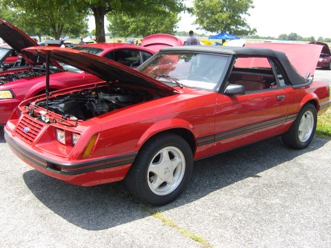 2007 Ford Mustang - Red convertible Mustang parked at an outdoor car show, hood up, next to another red Mustang.