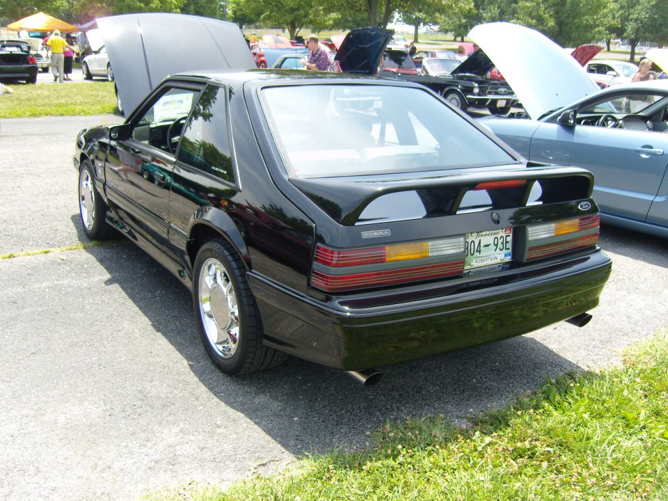 Black coupe with hood up, rear view showing spoiler and license plate. Other cars and tents in background