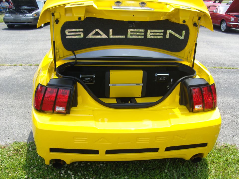 Yellow Saleen Mustang rear view with trunk open, showing custom audio equipment and 'SALEEN' badging on the trunk lid and bumper