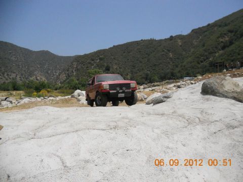 1988 Ford Bronco - A red SUV navigates a rocky, off-road path under a clear blue sky.