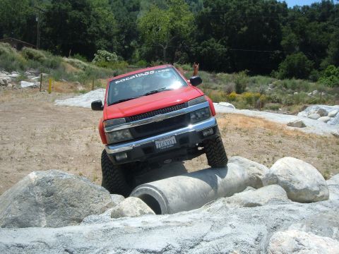 1988 Ford Bronco - A red pickup truck navigates a rocky off-road course, with its front tires on a concrete pipe.
