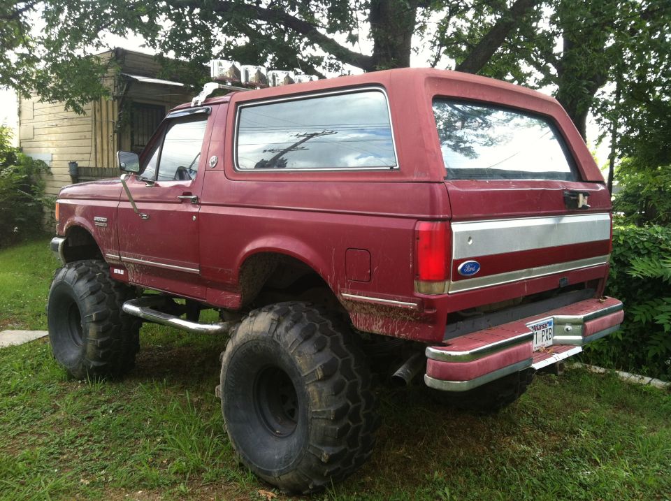 Red lifted Ford Bronco with large off-road tires, chrome side steps, and a silver stripe on the tailgate
