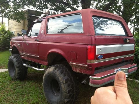 1991 Ford Bronco - This lifted Ford Bronco has massive tires and a custom rear bumper, sitting in a grassy yard.