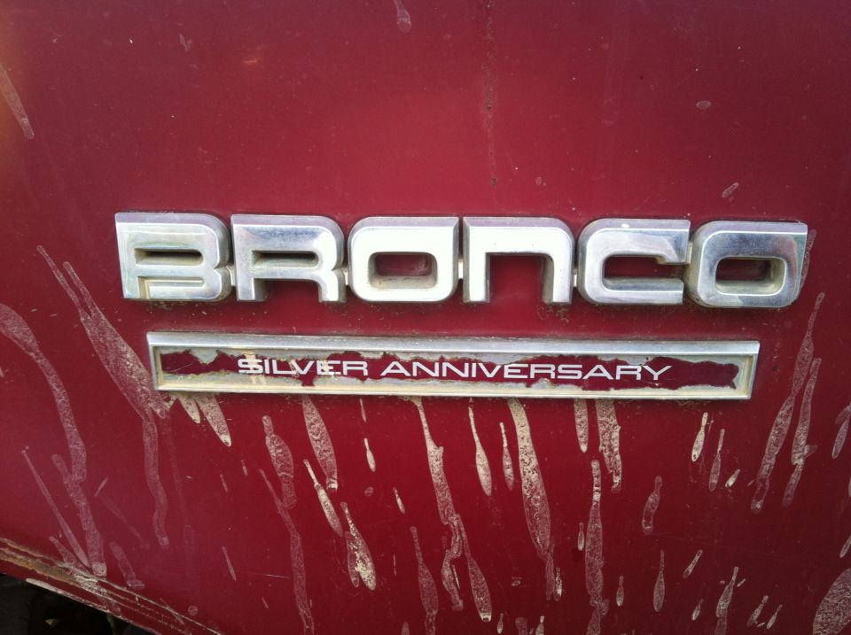 Red vehicle panel with chrome 'BRONCO' emblem and 'SILVER ANNIVERSARY' badge below, showing dirt streaks