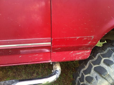 1991 Ford Bronco - Close-up of a red vehicle's side panel, showing chrome trim, a large tire, and numerous scratches.