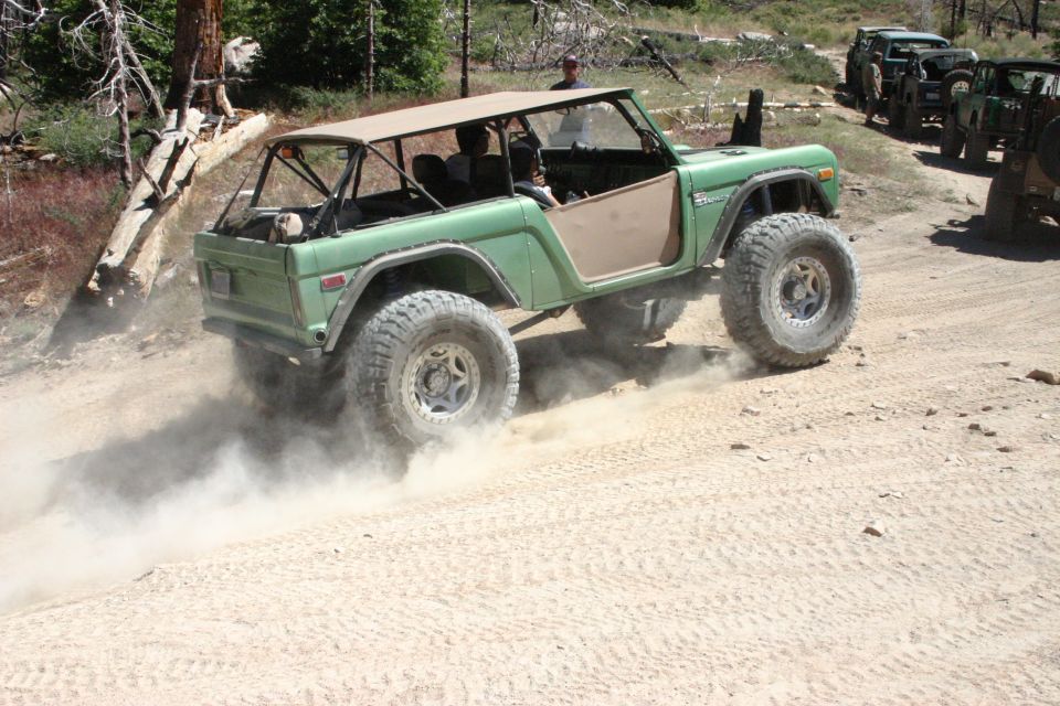 Green Ford Bronco with a tan soft top driving on a dirt road, kicking up dust