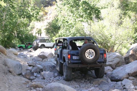 1991 Ford Bronco - Several off-road vehicles navigate a boulder-strewn path through a wooded canyon.