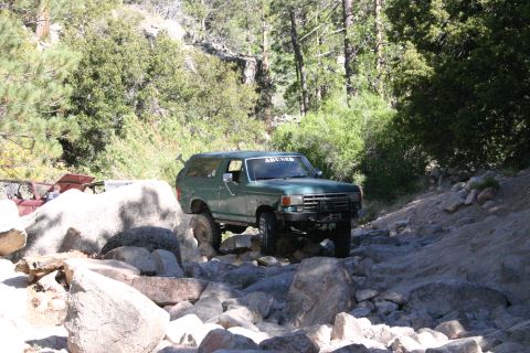 1991 Ford Bronco - This green SUV, likely a 1987 Ford Bronco, tackles a challenging rocky trail with "ABUSED" visible on its windshield.