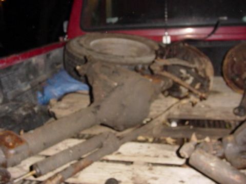 1992 Ford Bronco - Rear axle parts and a spare tire are loaded into the bed of a red pickup truck.