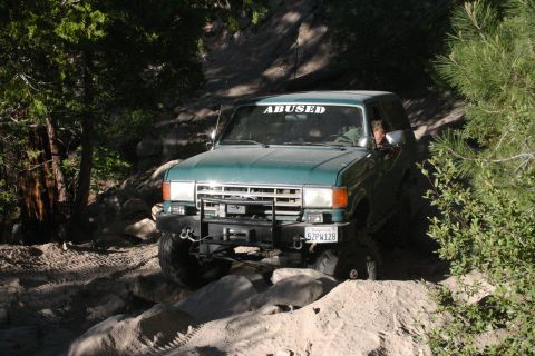 1991 Ford Bronco - This SUV is navigating a rocky off-road trail with trees on either side.
