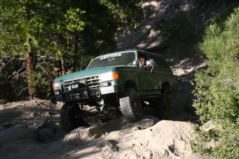 1991 Ford Bronco - A green Ford Bronco navigates a challenging off-road path with rocky terrain and dense foliage.