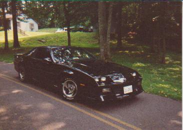 1982 Ford Bronco - A black sports coupe is parked on the side of a road with trees and a house in the background.