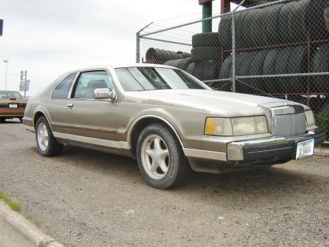 1987 Lincoln Mark VII - A tan 1987 Lincoln Mark VII coupe is parked outdoors, with a stack of tires visible behind a fence.