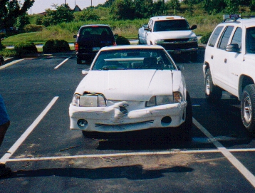 1982 Ford Bronco - A white sedan with significant front-end damage is parked in a lot with other vehicles.
