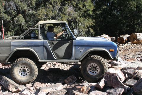 1991 Ford Bronco - A blue 1991 Ford Bronco is shown from the side, driving over a rocky terrain with trees in the background. The vehicle has a soft top and t…