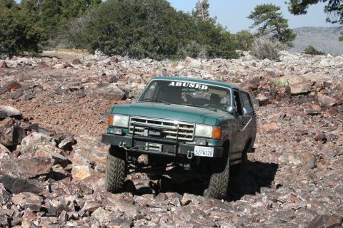 1991 Ford Bronco - A green Ford Bronco navigates a rocky, off-road terrain under a clear sky.
