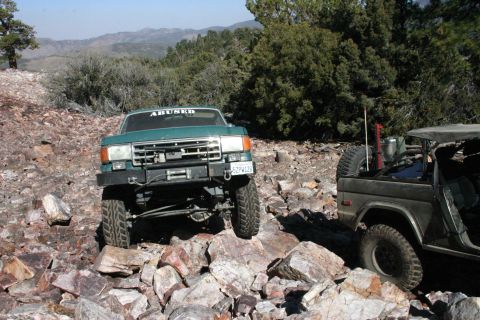 1991 Ford Bronco - Two Broncos tackle a rocky trail in a mountainous, wooded area under a clear blue sky.