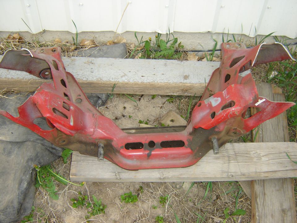 Red steel front crossmember subframe with rust, laid on wood boards outdoors