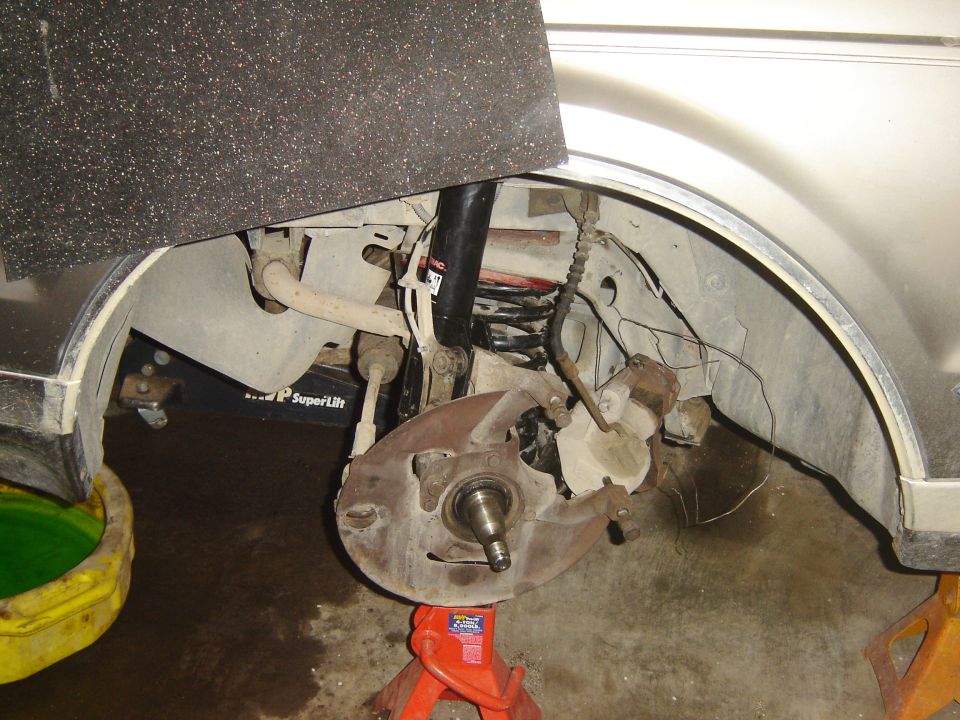 Front suspension with wheel and brake rotor removed, bare spindle and strut visible, car on jack stand in garage