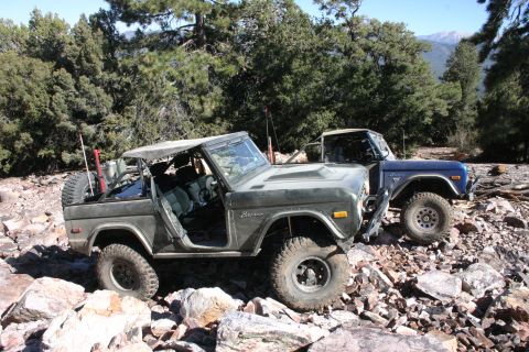 1991 Ford Bronco - Two Ford Broncos navigate a rocky off-road path in a wooded, mountainous area.