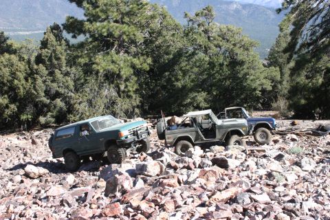 1991 Ford Bronco - Several SUVs navigate a challenging, rocky mountain path surrounded by trees and distant mountains.