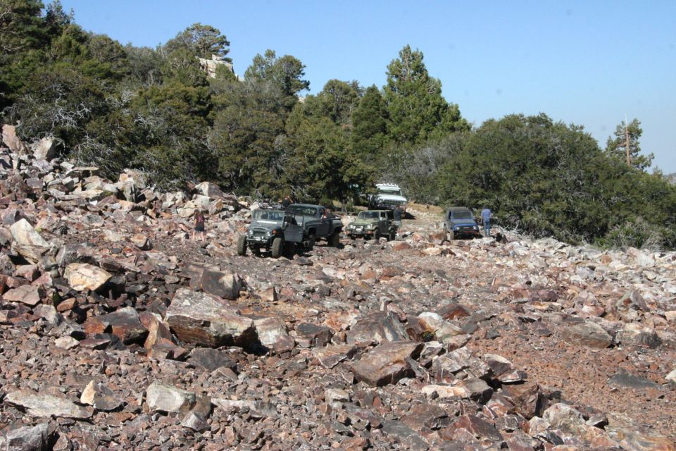 Several off-road vehicles navigate a steep, rocky trail with sparse trees and clear blue sky