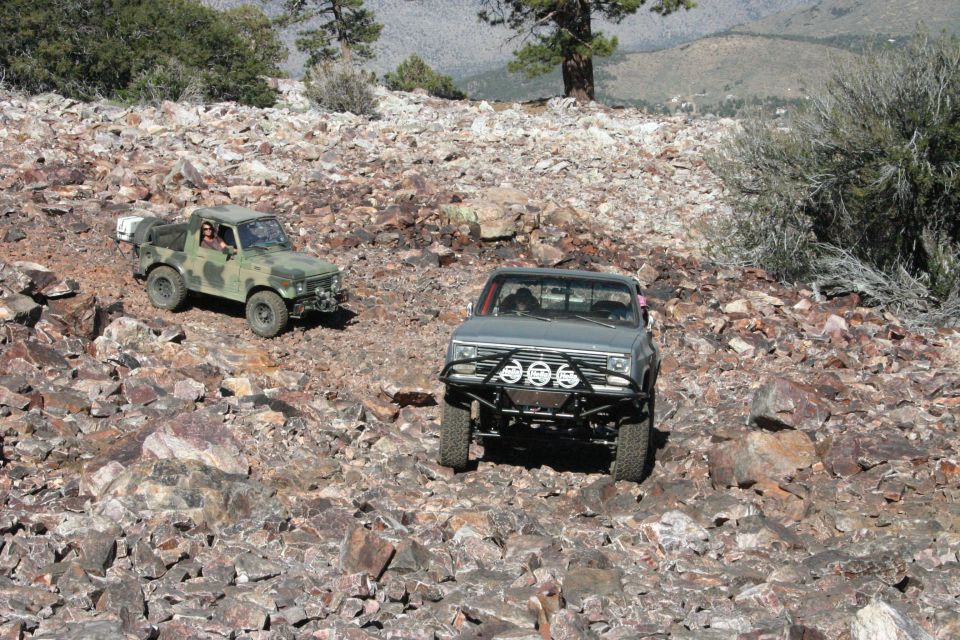 Two modified trucks are driving on a rocky, mountainous trail. One is a camouflage-colored pickup, the other is a gray pickup with a brush guard