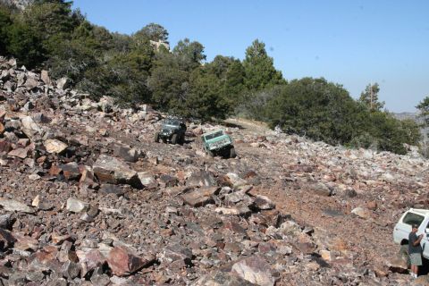 1991 Ford Bronco - Several off-road vehicles traverse a steep, rocky terrain with sparse trees and brush.