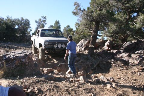 1991 Ford Bronco - A person walks near a modified pickup truck navigating a rocky off-road trail under a clear blue sky.