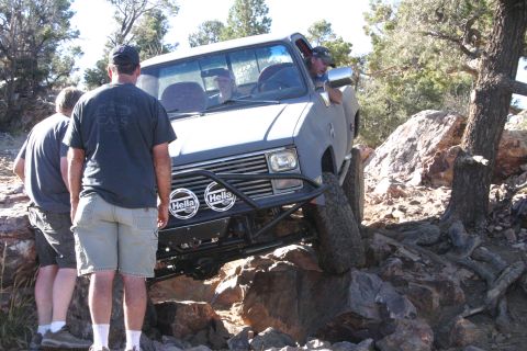 1991 Ford Bronco - A gray pickup truck is shown off-roading over a rocky obstacle with two people observing.