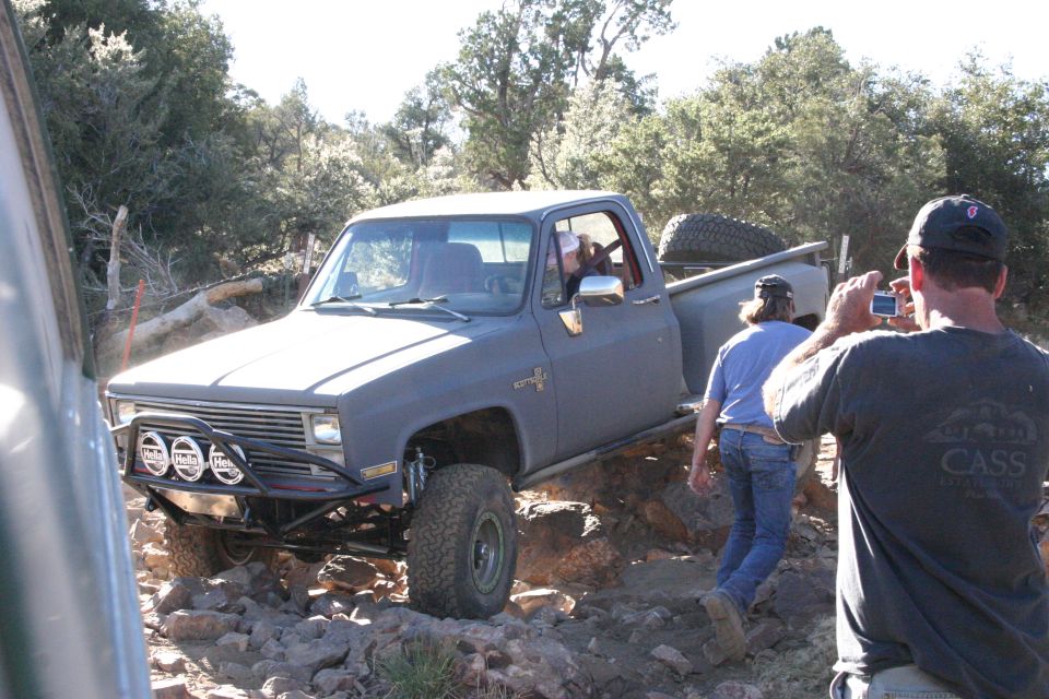 Gray pickup truck with off-road tires and lights navigating a rocky trail. A person is taking a photo of it