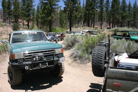 1991 Ford Bronco - A group of off-road vehicles are parked on a dirt road surrounded by trees and brush.