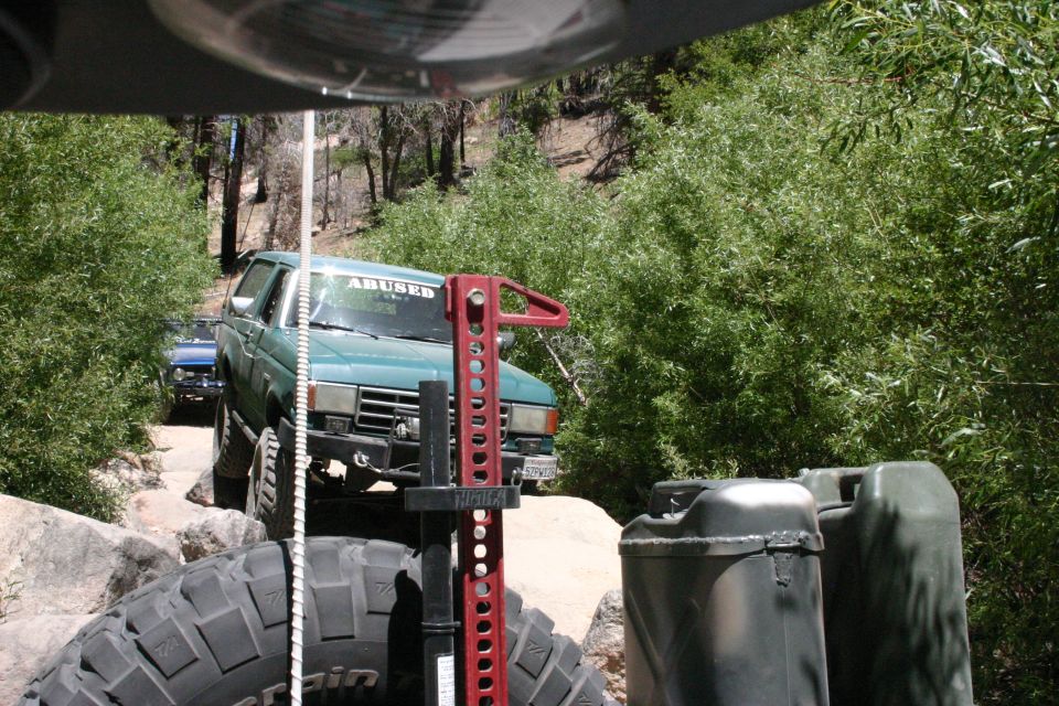 Green Ford Bronco on a rocky trail, with a blue vehicle behind it. A Hi-Lift jack and jerry cans are in the foreground