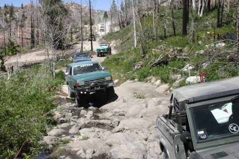 1991 Ford Bronco - Several off-road vehicles navigate a challenging rocky path through a wooded area.
