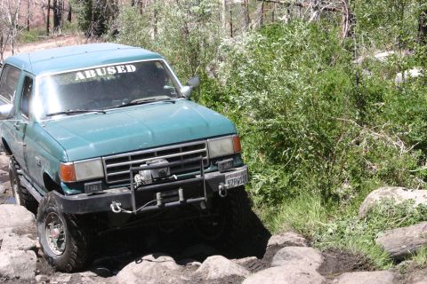 1991 Ford Bronco - This SUV is navigating a rocky trail with dense foliage on the right side.