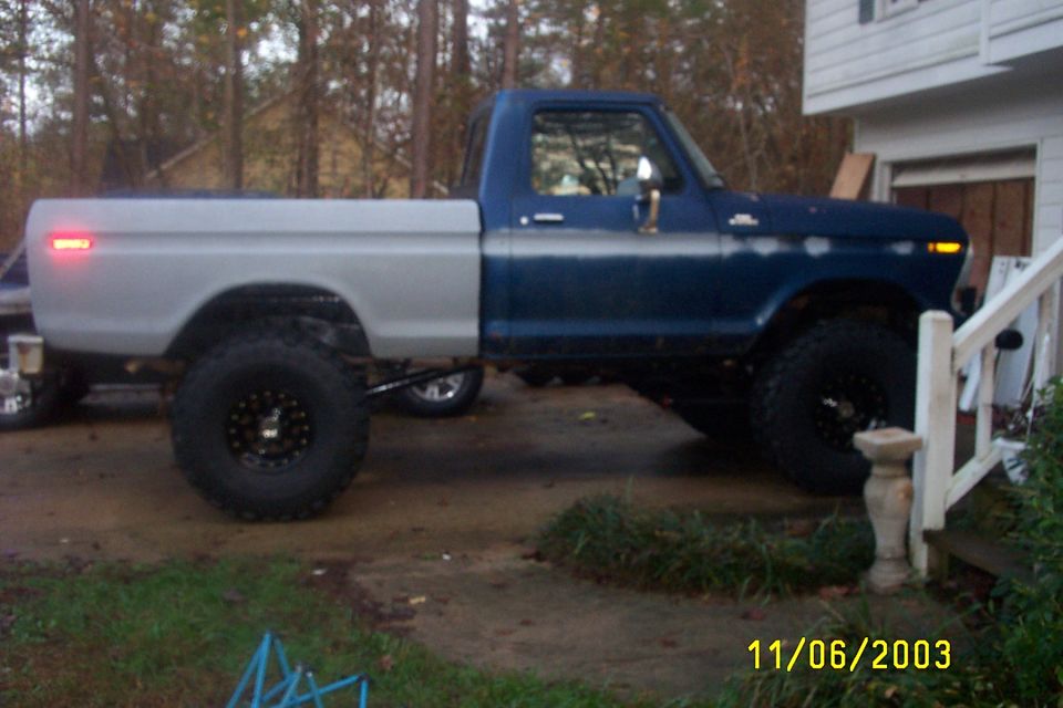 Lifted 1970s Ford F-150 with blue cab, primer-gray bed and large mud tires, side view in a driveway