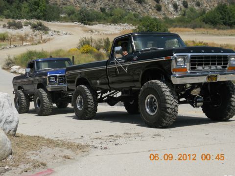 1986 Ford Bronco - Two lifted Ford pickup trucks are seen on a dirt road, with the black truck in the foreground.