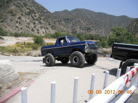 1986 Ford Bronco - A lifted blue pickup truck tows a black pickup truck on a road with a mountainous backdrop.
