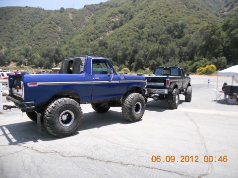 1986 Ford Bronco - A lifted blue Ford Bronco tows a black Ford pickup truck on a sunny day with a mountainous backdrop.