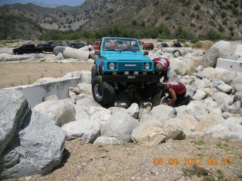 1986 Ford Bronco - Two people inspect a lifted blue Suzuki Samurai navigating a rocky terrain. Other vehicles are parked in the background.