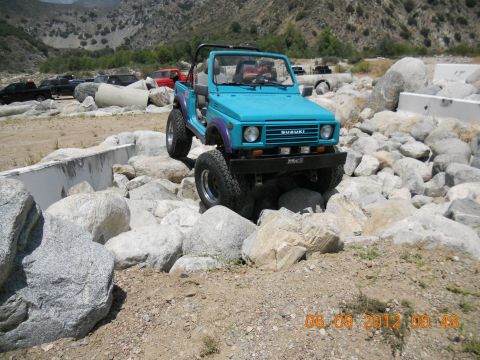 1986 Ford Bronco - A teal Suzuki Samurai navigates a rocky obstacle course with other vehicles in the background.